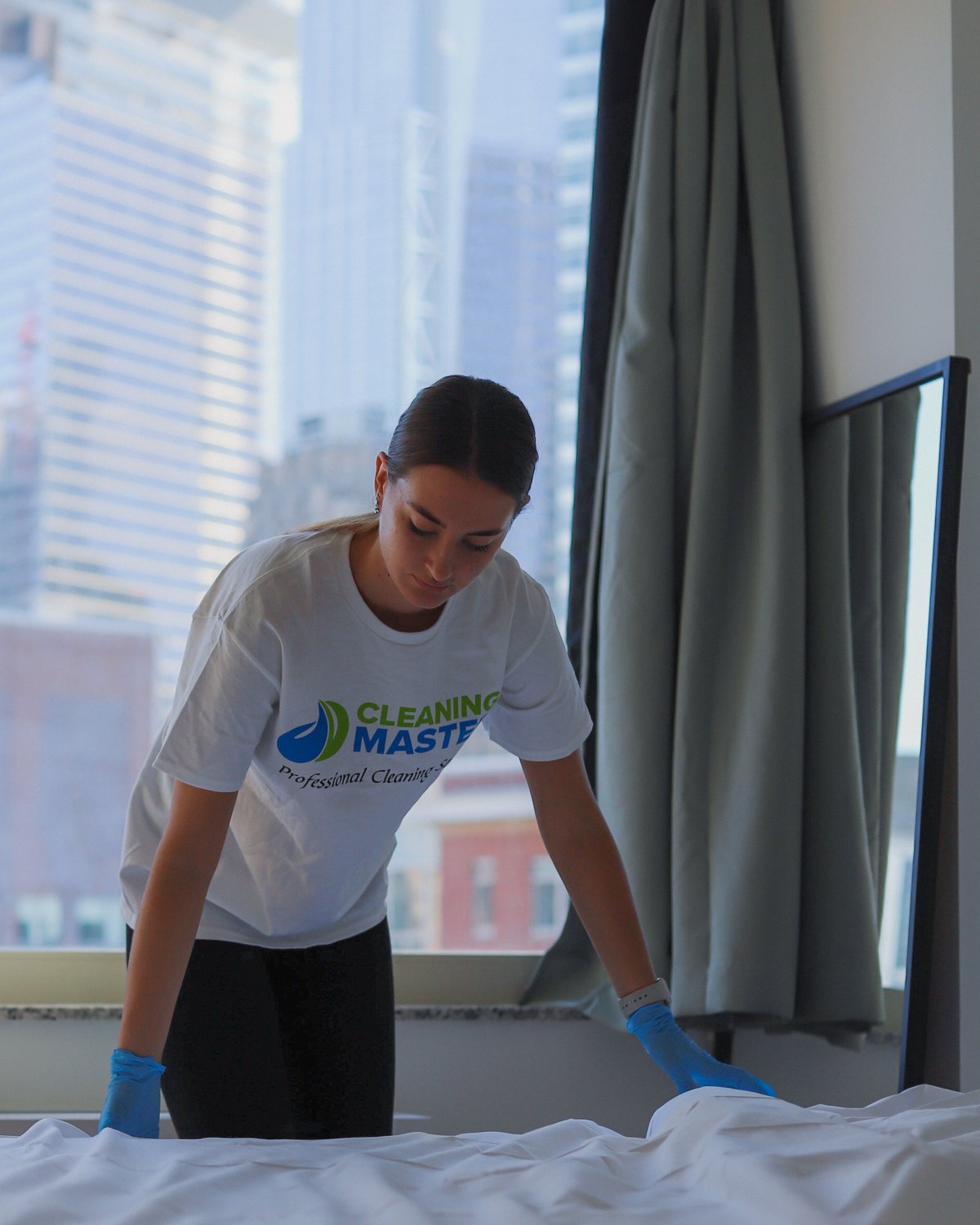Cleaning Master professional making a bed in a high-rise apartment with the New York skyline behind